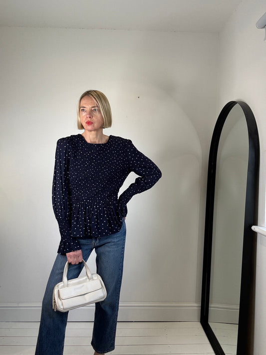 Woman in a navy blue polka dot blouse and jeans holding a white handbag, standing next to a tall mirror.